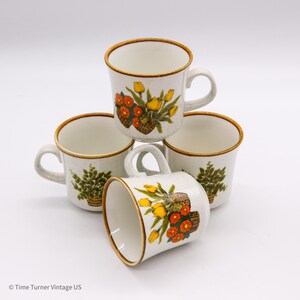 May include: Four vintage ceramic mugs with a speckled cream background. Each mug features a brown rim and handle, and is decorated with a floral design of yellow and orange flowers in a basket. One mug has a fern design.