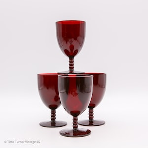May include: Four ruby red wine glasses. The glasses are arranged in a pyramid shape, with one glass balanced on top of the other three. Each glass has a rounded bowl and a textured stem.