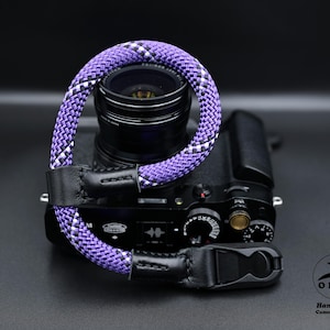 May include: A black camera with a purple and white rope camera strap. The strap is attached to the camera with black leather ends and a black plastic buckle. The camera lens is visible.
