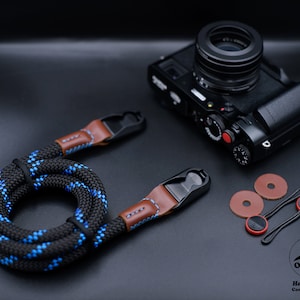 May include: A black and blue camera strap with brown leather accents and metal clips. The strap is coiled up and the camera is not attached. The strap is made by OKLI Handmade Camera Straps.
