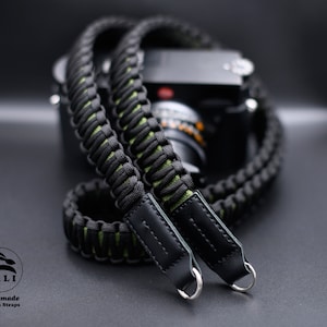 May include: A black and green camera strap with leather ends and metal rings. The strap is made of paracord and has a braided design. The strap is handmade and is a great way to add a touch of style to your camera.