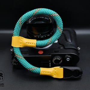 May include: A teal, pink, and yellow camera strap with a leather strap and a metal clasp. The strap is attached to a black camera.
