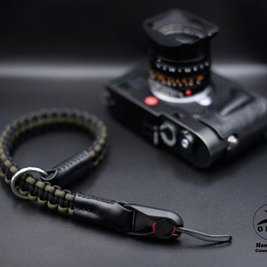 Könnte beinhalten: Eine schwarze und olivgrüne Paracord-Kamerahandschlaufe mit schwarzem Lederakzent und einem silbernen Metallring. Im Hintergrund befindet sich eine schwarze Kamera. Der Gurt ist handgefertigt.