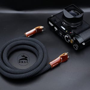 Op de afbeelding: Een zwarte camerariem van gevlochten touw met bruine lederen uiteinden, witte kruissteken en zilverkleurige metalen ringen. De riem ligt opgerold naast een zwarte digitale camera. Een rond logo met een konijn en de tekst OKLI is zichtbaar.