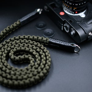 Puede incluir: Correa de cámara de paracord verde oliva con detalles de cuero negro y anillos metálicos. La correa está enrollada sobre una superficie oscura junto a una cámara negra con una funda de cuero. El logotipo "OKLI Handmade Camera Straps" es visible.