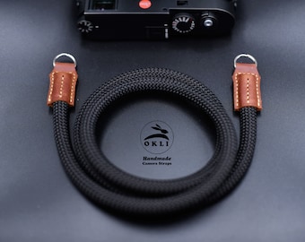 Könnte beinhalten: Schwarzer Kameragurt mit braunen Lederenden und silbernen Metallringen. Der Gurt ist aus geflochtenem Seil und trägt den Text "OKLI Handmade Camera Straps".