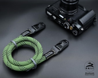 Op de afbeelding: Een groene en zwarte cameraband met een leren accent en een zwarte metalen gesp. De band is opgerold en klaar om aan een camera te worden bevestigd.