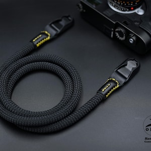 Könnte beinhalten: Ein schwarzer Kameragurt aus geflochtenem Seilmaterial. Der Gurt hat schwarze Lederenden mit gelben Nähten. Im Hintergrund ist eine schwarze Kamera zu sehen. Der Gurt stammt von OKLI Handmade Camera Straps.
