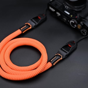 May include: A bright orange braided rope camera strap featuring black leather connectors with orange stitching and black plastic quick-release buckles with red buttons, attached to a black digital camera body.