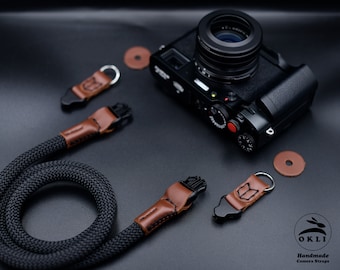 Op de afbeelding: Zwarte cameraband met bruine lederen accenten en metalen hardware. De band is opgerold en ligt op een zwart oppervlak met een camera en andere accessoires.