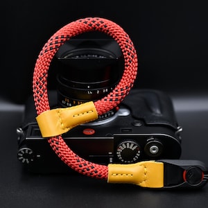 Può includere: Un cinturino da polso per fotocamera con cordino intrecciato rosso e nero, caratterizzato da inserti e cuciture in pelle gialla. Il cinturino è fissato a un corpo macchina nero con obiettivo nero, mostrando un design ad anello.