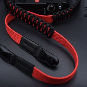 May include: Red and black camera strap with black leather accents and metal clasps. The strap is made of paracord and has a woven design. The strap is labelled "OKLI Handmade Camera Straps".