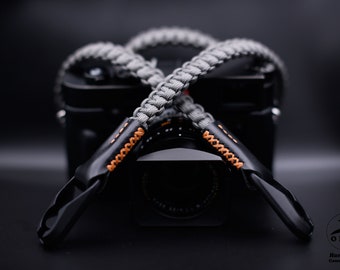 Op de afbeelding: Een grijze gevlochten camerariem met zwarte leren uiteinden en oranje stiksels. De riem ligt over een zwarte camerabody, met de lens zichtbaar. Het is een handgemaakt product.