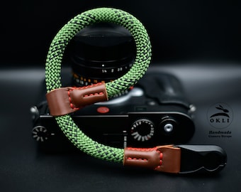 Könnte beinhalten: Ein grünes und schwarzes Kameraband mit roten Nähten und braunen Lederakzenten. Das Band ist an einer Kamera mit einem schwarzen Objektiv befestigt. Das Band wird von OKLI Handmade Camera Straps hergestellt.