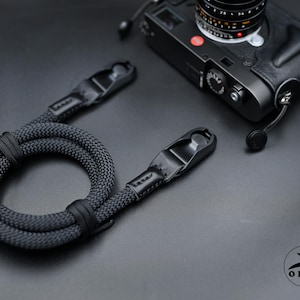 May include: A black rope camera strap with textured braided material and smooth black leather connectors at both ends. The strap is coiled next to a black camera body. A circular logo with a rabbit silhouette and the text OKLI is visible.