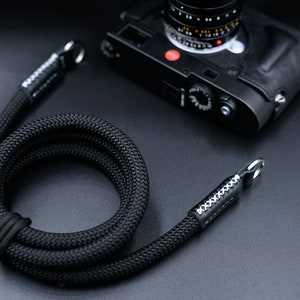 May include: Black camera strap made of braided rope with leather ends and silver hardware. The strap is coiled and laid on a black surface. The brand name "OKLI Handmade Camera Straps" is visible in the bottom right corner.