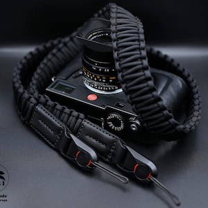 May include: Black camera strap made of paracord with a leather pad and a metal clip. The strap is attached to a black camera with a lens.