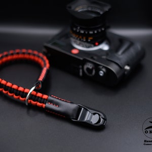 May include: A black and red woven camera strap with a black leather end and metal fittings. A black camera is blurred in the background. The strap features the text "OKLI Handmade Camera Straps".