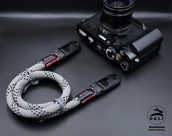 Op de afbeelding: Een grijs en zwart patroon camerariem met roze stiksels en zwarte lederen uiteinden. De riem is opgerold naast een zwarte camera. Het logo "OKLI Handmade Camera Straps" is zichtbaar.