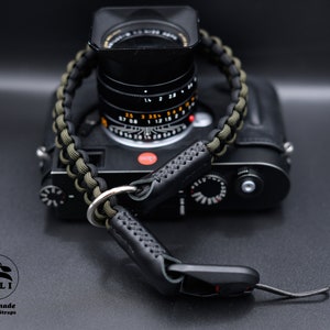 May include: A black camera with a braided olive green and black strap. The strap has black leather ends and silver metal rings. The camera strap is handmade. The logo says "OKLI Handmade Camera Straps".