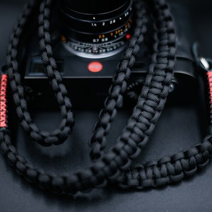 May include: Black braided camera strap with red stitching. The strap is made of a thick, durable material and is designed to be worn around the neck. The camera strap is shown on a black camera.