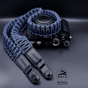 May include: A blue paracord camera strap with leather accents and a black camera attached. The strap is handmade and features the text "OKLI Handmade Camera Straps".