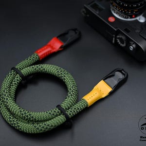 Puede incluir: Correa de cámara con estampado verde y negro con detalles de cuero rojo y amarillo. La correa está hecha de un material tejido duradero y presenta conectores negros. También se ve una cámara negra.