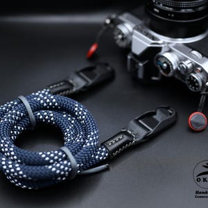 May include: A navy blue and white speckled camera strap with black leather ends. The strap is coiled on a black surface near a silver and black camera. The strap is handmade.