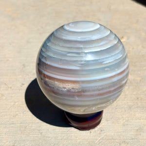 May include: A polished, spherical stone orb featuring horizontal bands of white, gray, and translucent brown layers. The orb is mounted on a small, dark reddish-brown circular wooden stand, showcasing natural mineral banding and a smooth, glossy finish.