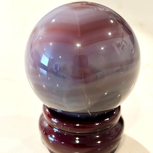 May include: A polished, spherical purple agate stone with visible horizontal banding and a smooth, reflective surface, resting securely on a dark, circular wooden display stand that features a glossy finish and a raised, rounded edge design.