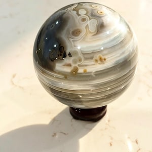 May include: A polished spherical stone featuring intricate banding in shades of gray, white, and translucent brown. The surface displays circular eye-like patterns and dark inclusions, resting on a small, dark, circular base that supports the sphere.