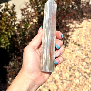 May include: A tall, hexagonal crystal tower with a pointed top, featuring vertical bands of translucent white and clear mineral inclusions. The stone has a smooth, polished surface that reflects light, displaying a columnar structure throughout its length.