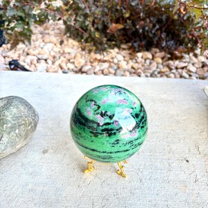 May include: A polished spherical gemstone featuring a vibrant green base with prominent black horizontal bands and scattered magenta-colored inclusions, resting securely on a small, three-legged gold-toned metal display stand.