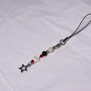 May include: A beaded phone charm with a star charm. The charm features a silver star, white, black, and red beads, and a white star-shaped charm. It has a black cord and a silver clasp.