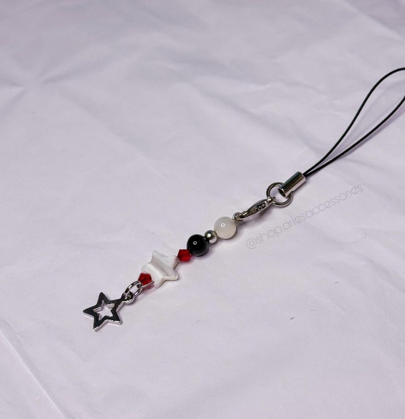 May include: A beaded phone charm with a star charm. The charm features a silver star, white, black, and red beads, and a white star-shaped charm. It has a black cord and a silver clasp.