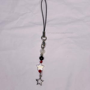 May include: A beaded phone charm with a black cord and silver-tone hardware. The charm features white, black, and red beads, two star-shaped charms, and a hollow star pendant. The text "@shop.ariesaccessories" is visible.