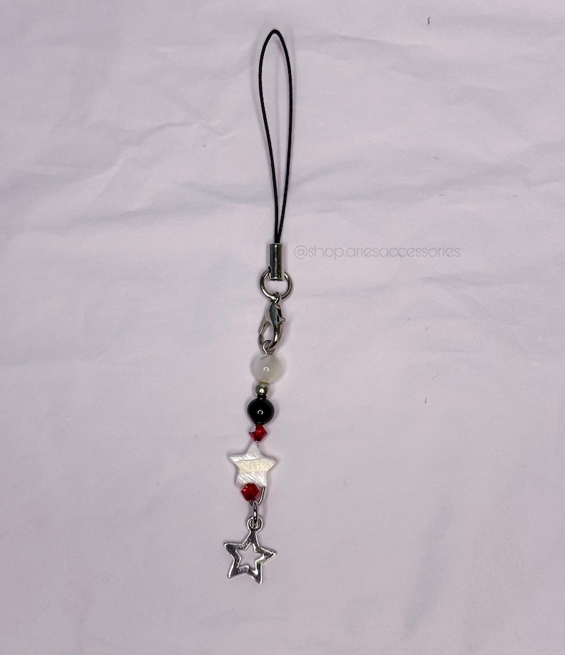 May include: A beaded phone charm with a black cord and silver-tone hardware. The charm features white, black, and red beads, two star-shaped charms, and a hollow star pendant. The text "@shop.ariesaccessories" is visible.