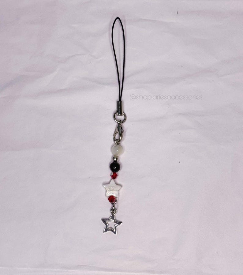 May include: A beaded phone charm with a black cord, silver clasp, and a dangling star charm. The charm features white, black, and red beads, with the text "@shop.ariesaccessories".