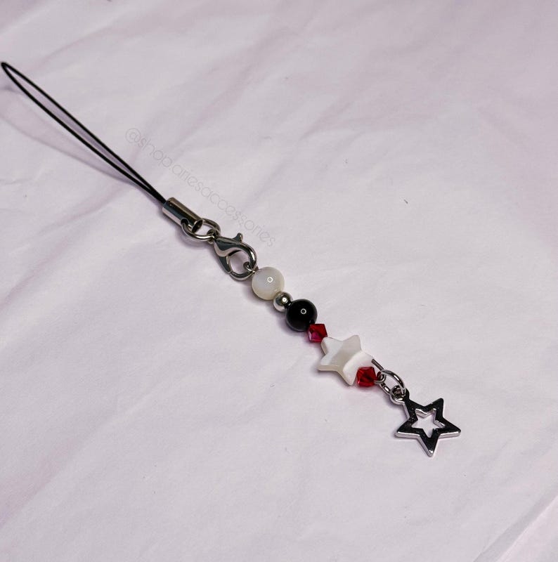 May include: A beaded phone charm with a black cord and silver-tone clasp. The charm features white, black, and red beads, along with a white star-shaped bead and a silver star charm. The text "@shopariesaccessories" is visible.