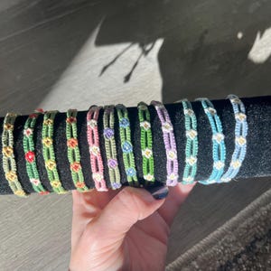 May include: A collection of colorful macrame bracelets. Each bracelet features a woven design with small floral accents in various colors, including red, yellow, pink, and blue. The bracelets are displayed on a black surface.