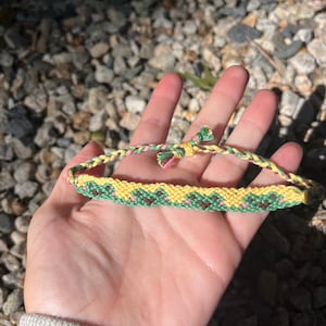 May include: A green, yellow, and pink friendship bracelet with a pattern of hearts. The bracelet is made with embroidery floss and is tied in a knot.