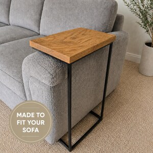Pu&ograve; includere: Un tavolino in legno con piano rettangolare e struttura in metallo nero. Il piano ha una finitura a venatura naturale. Il testo "MADE TO FIT YOUR SOFA" &egrave; in un cerchio. Progettato per divani.