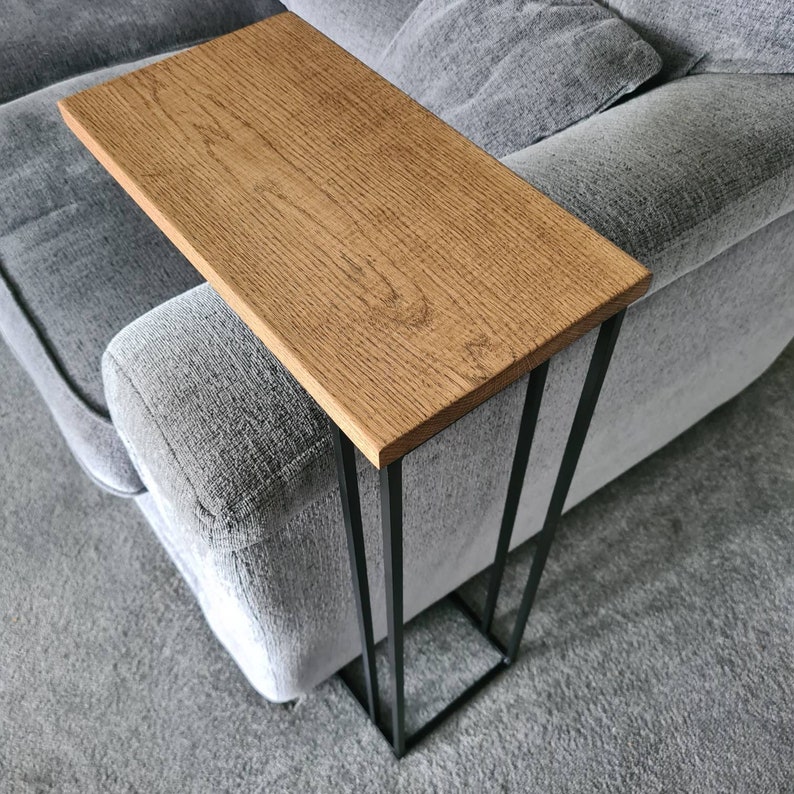 May include: A wooden side table with a rectangular oak top and a black metal frame. The table is designed to slide over the arm of a sofa. The wood grain is visible on the table's surface, and the frame has a minimalist design.