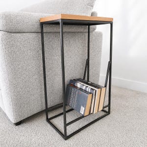 May include: A contemporary side table featuring a black metal frame and a wooden top. The table includes a book rack holding several books. It is positioned beside a grey sofa on a light grey carpet.
