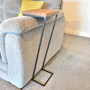 May include: A side table with a rectangular, light brown wooden top and a black metal frame. The frame has a unique, angled design, with a square base. The table is placed next to a grey sofa.