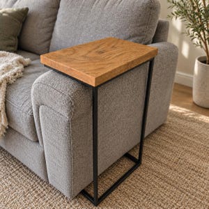 May include: A C-shaped side table featuring a rectangular natural wood grain top and a slim black metal frame. The table is designed to slide over the arm of a sofa, providing a flat surface for items. The construction consists of a solid wood tabletop supported by a continuous, thin, rectangular black metal base that rests on the floor.