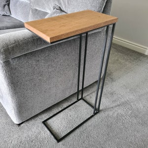May include: A side table with a rectangular, light brown wooden top and a black metal frame. The table is designed to slide alongside a sofa. The table's dimensions are approximately 24 inches long, 12 inches wide, and 24 inches high.