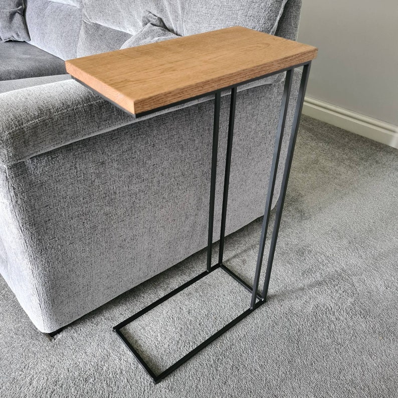May include: A side table with a rectangular, light brown wooden top and a black metal frame. The table is designed to slide alongside a sofa. The table's dimensions are approximately 24 inches long, 12 inches wide, and 24 inches high.