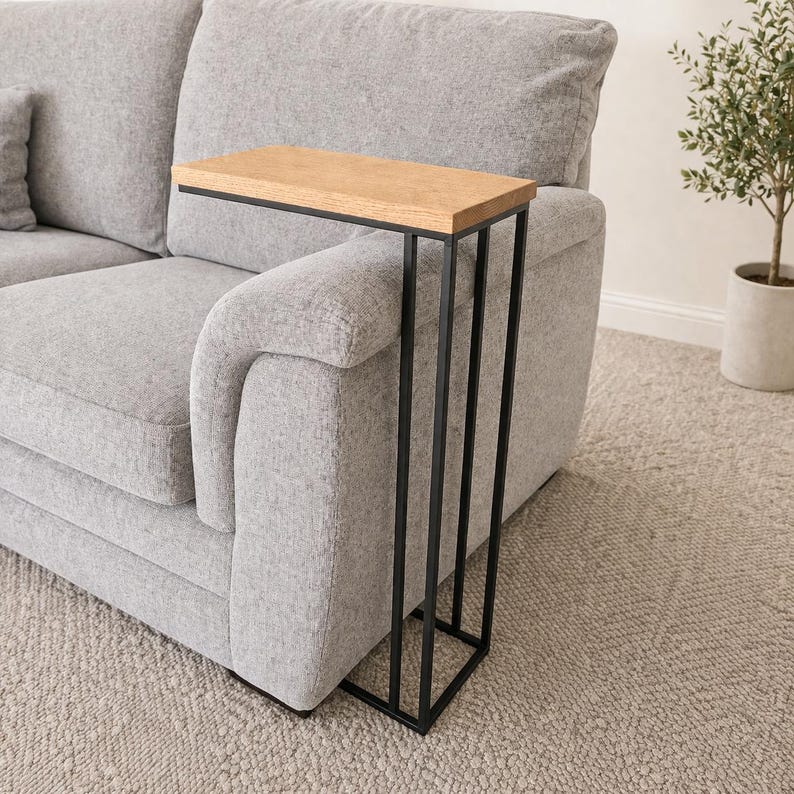 May include: A side table with a rectangular light wood top and a black metal frame. The table is designed to slide over the arm of a gray sofa. The table's dimensions are not visible in the image.