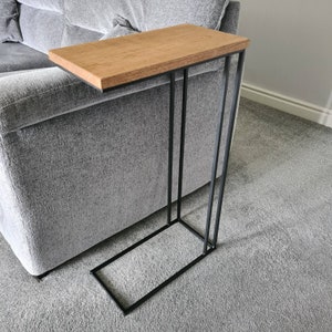 May include: A modern side table with a rectangular, light brown wooden top and a black metal frame. The table is designed to slide alongside a sofa. The table's dimensions are approximately 24 inches long, 12 inches wide, and 24 inches high.